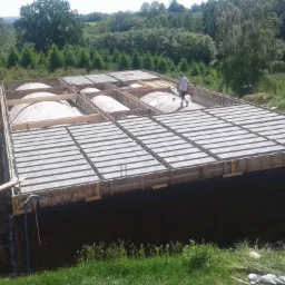 Budowa fundamentów domu z widoczną konstrukcją szalunkową, izolacją pionową i pracownikiem na górze, w tle zielony krajobraz.