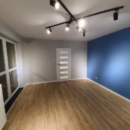 Pomalowane wnętrze pokoju z panelami podłogowymi, niebieską ścianą i oświetleniem szynowym.