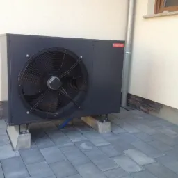 Ciemnoszara jednostka zewnętrzna z wentylatorem, zamontowana na betonowych podstawach na szarej kostce brukowej, z widocznym odpływem skroplin i fragmentem elewacji budynku z orynnowaniem.