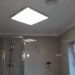 Wykończona łazienka z kabiną prysznicową, płytkami w odcieniach szarości, oświetleniem sufitowym LED i wentylatorem.