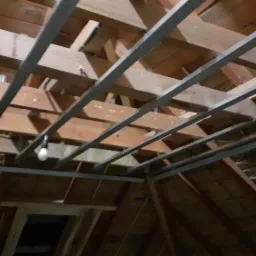 Konstrukcja metalowa podwieszana do drewnianego stropu poddasza, widoczne elementy oświetlenia, przygotowanie do montażu płyt.