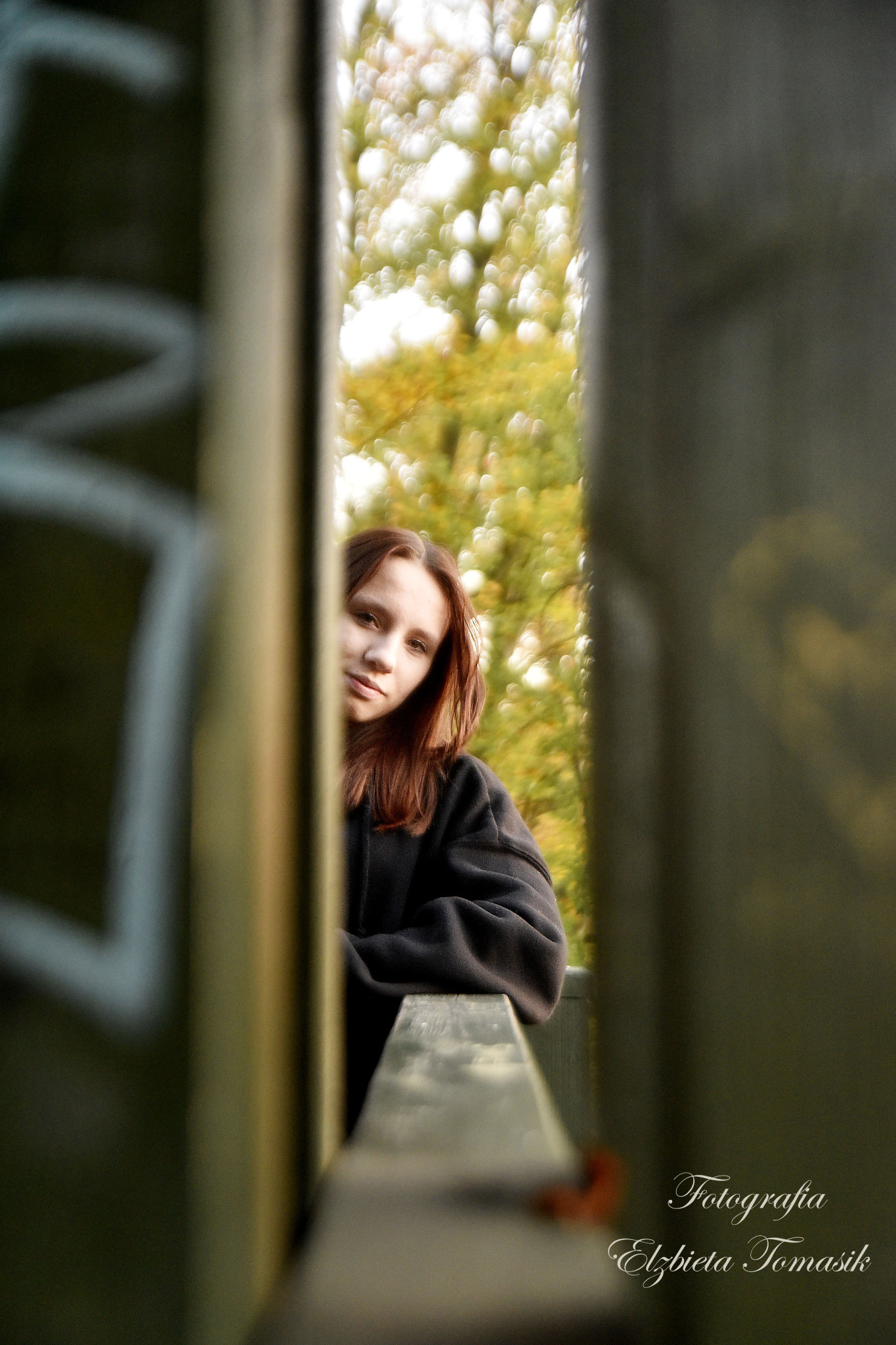 Portret kobiety z rudymi włosami, spoglądającej przez wąskie przejście w drewnianej konstrukcji, z rozmyte tłem drzew i jesiennych liści.