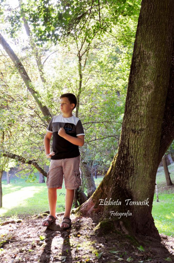 Chłopiec w koszulce w paski i krótkich spodenkach w kratę stoi oparty o duże drzewo w lesie, spoglądając w bok.