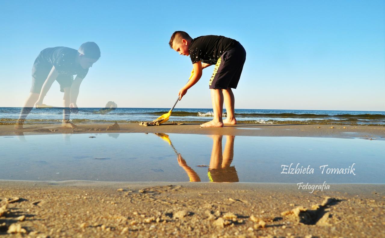 Chłopiec na plaży sprząta piasek żółtą łopatką, odbicie w wodzie, w tle morze i sylwetka osoby.