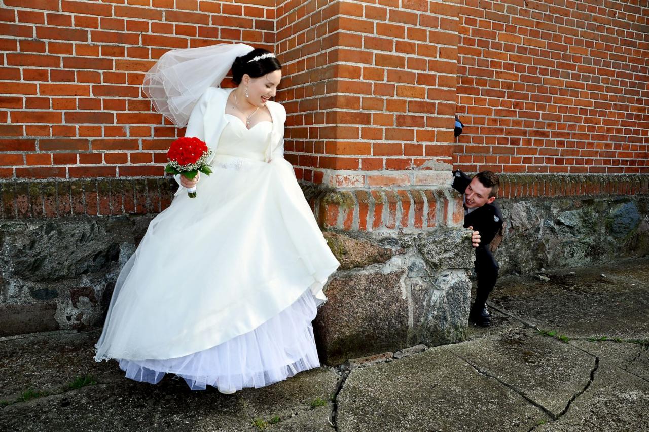 Panna młoda w białej sukni z bukietem czerwonych róż spogląda w dół, a pan młody w czarnym garniturze wygląda zza rogu ceglanej ściany.