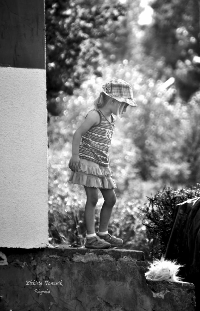 Czarno-białe zdjęcie dziewczynki w kapeluszu i sukience, stojącej na betonowej krawędzi z bujną roślinnością w tle, wykonane z niskiego kąta.