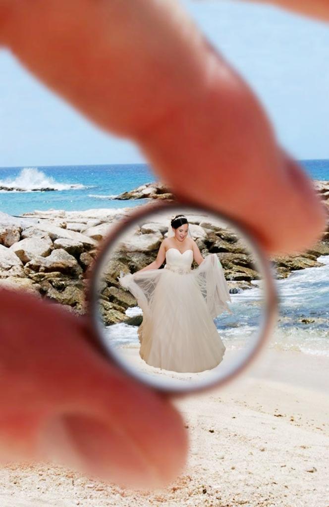 Panna młoda w białej sukni na plaży, widziana przez pierścień utworzony z palców, w tle morze i skały.