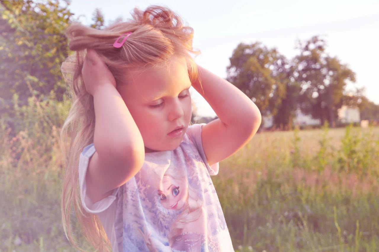 Dziewczynka z blond włosami i różową spinką, ubrana w koszulkę z Elsą, stoi na łące w promieniach zachodzącego słońca, trzymając ręce na głowie.