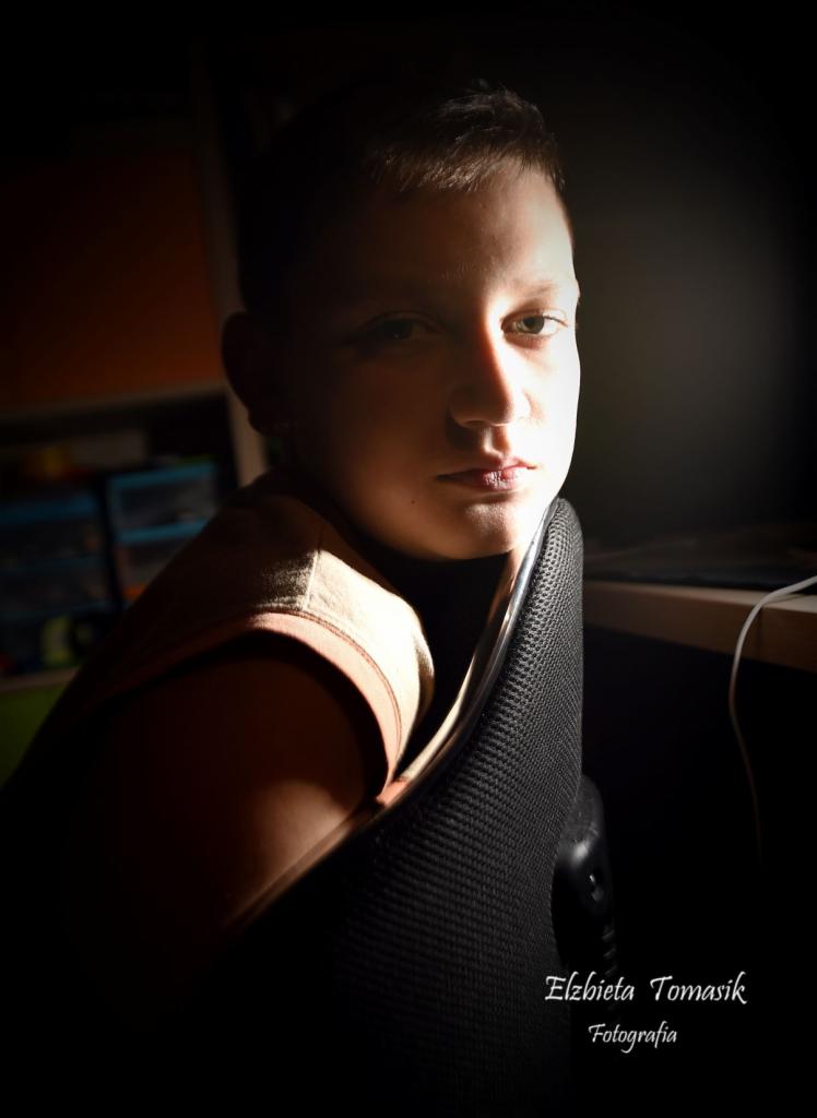 Portret młodego chłopca siedzącego na czarnym krześle biurowym, oświetlonego bocznym światłem, z widocznym fragmentem biurka i półki w tle.