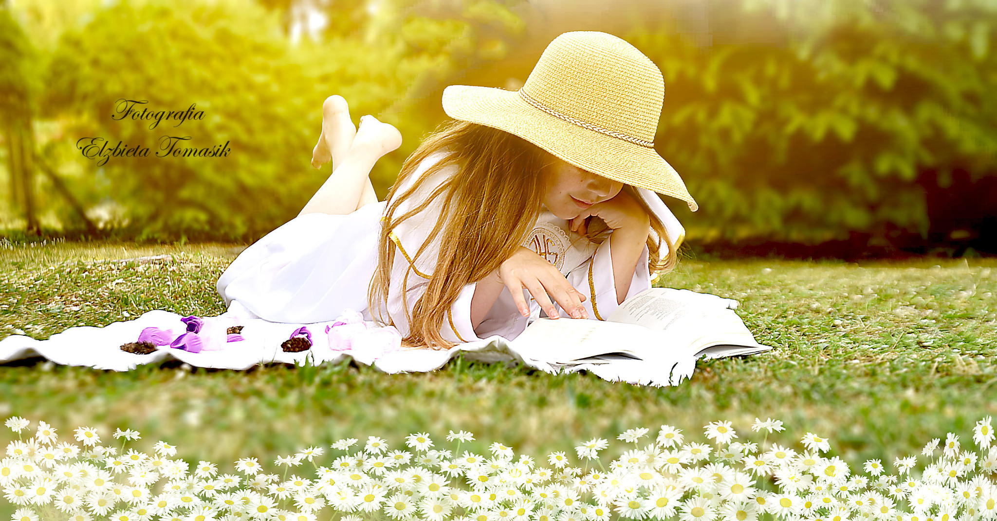 Dziewczynka w białej sukience i słomkowym kapeluszu leży na kocu w trawie, czytając książkę. W tle rozmyte drzewa i kwiaty, w dolnej części kadru stokrotki.