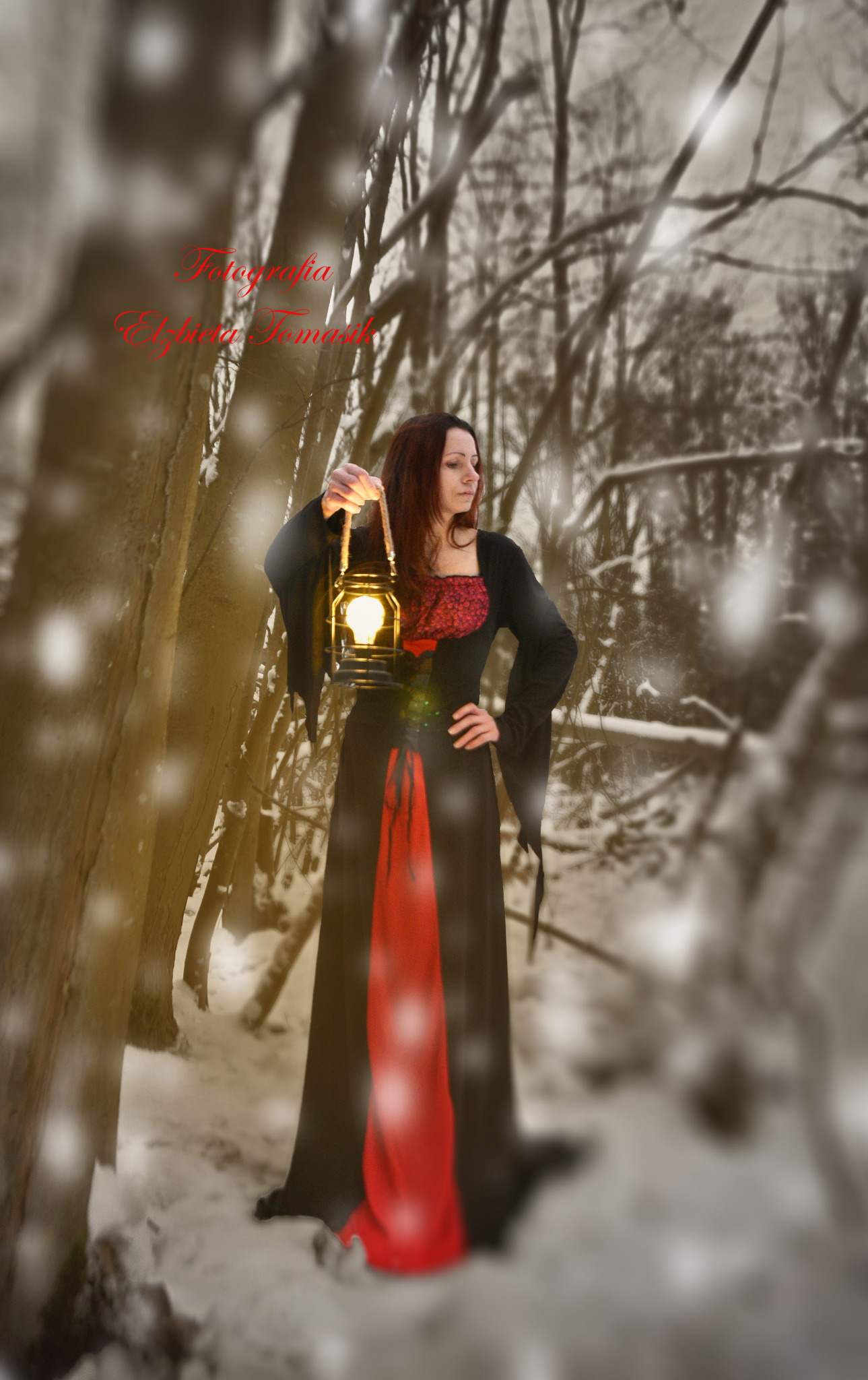 Kobieta w czarnej sukni z czerwonymi akcentami, trzymająca zapaloną latarnię w zimowym lesie.