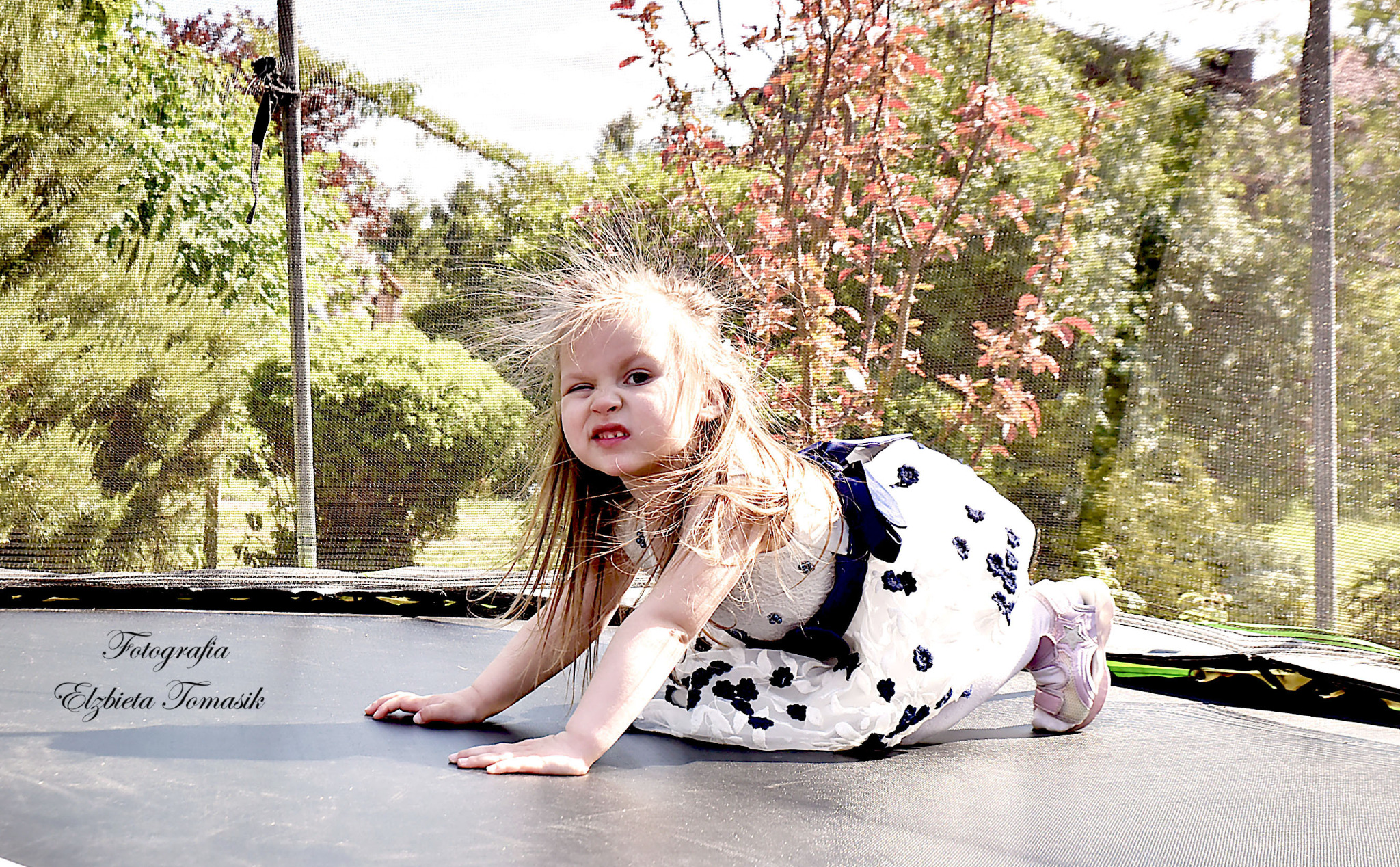 Dziewczynka w białej sukience z ciemnymi kwiatami, leżąca na trampolinie w ogrodzie, z włosami uniesionymi przez elektryczność statyczną i przymrużonym okiem.