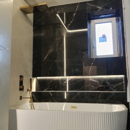 Voytex , Sufity Napinane , Wykonczenia , remonty - Nowoczesna łazienka z czarną marmurową ścianą, podświetleniem LED i białą wanną z żłobieniami. Złota armatura i deszczownica dodają elegancji.