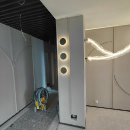 Voytex , Sufity Napinane , Wykonczenia , remonty - Nowoczesny korytarz z panelami ściennymi w odcieniach beżu i szarości, ozdobionymi geometrycznymi łukami. Trzy okrągłe kinkiety i czarny sufit lamelowy. Widoczne narzędzia i kable.