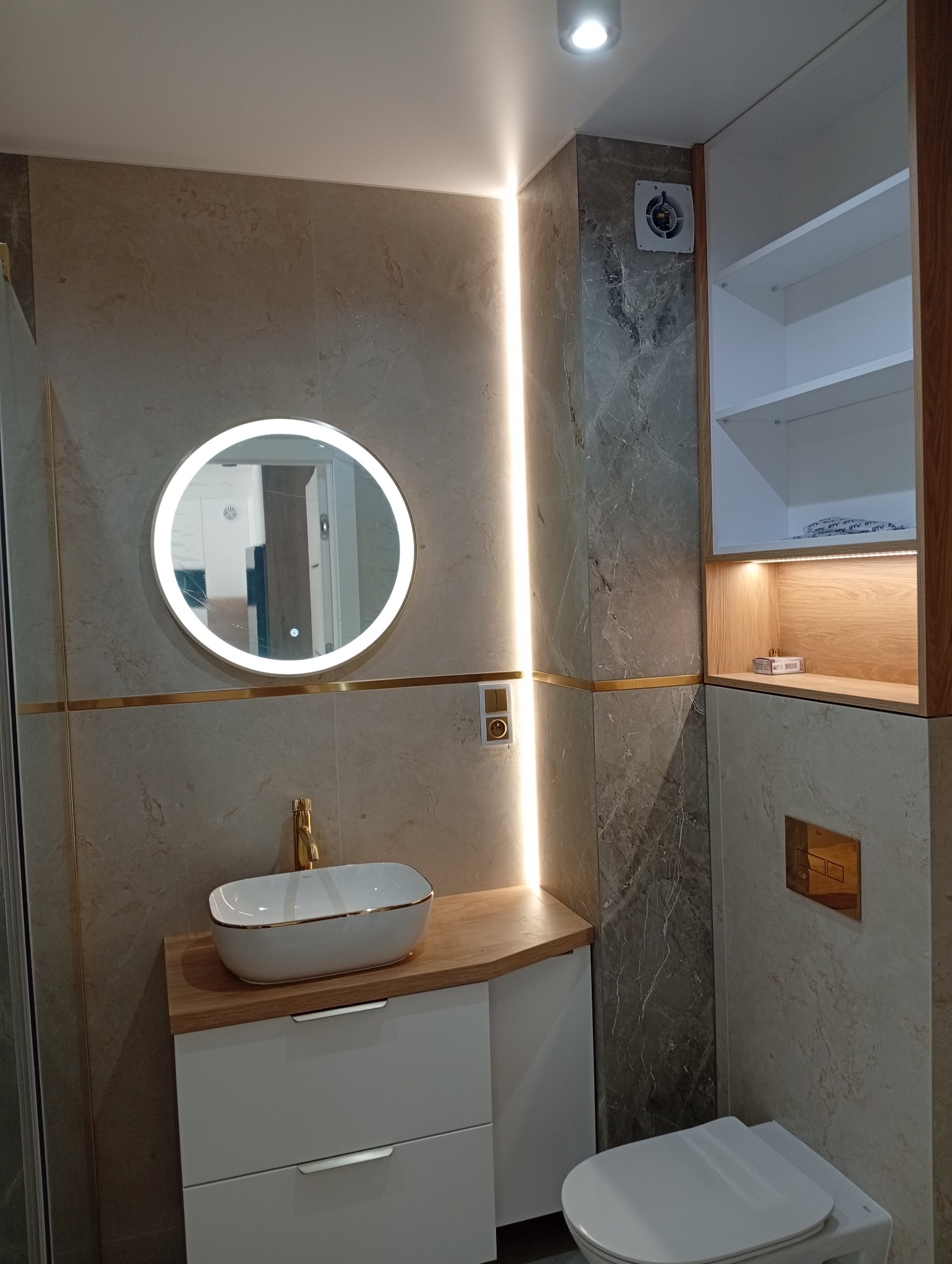 Nowoczesna łazienka z okrągłym lustrem LED, umywalką nablatową z złotymi akcentami i szafką. Płytki imitujące kamień i oświetlenie ledowe tworzą elegancki wystrój.