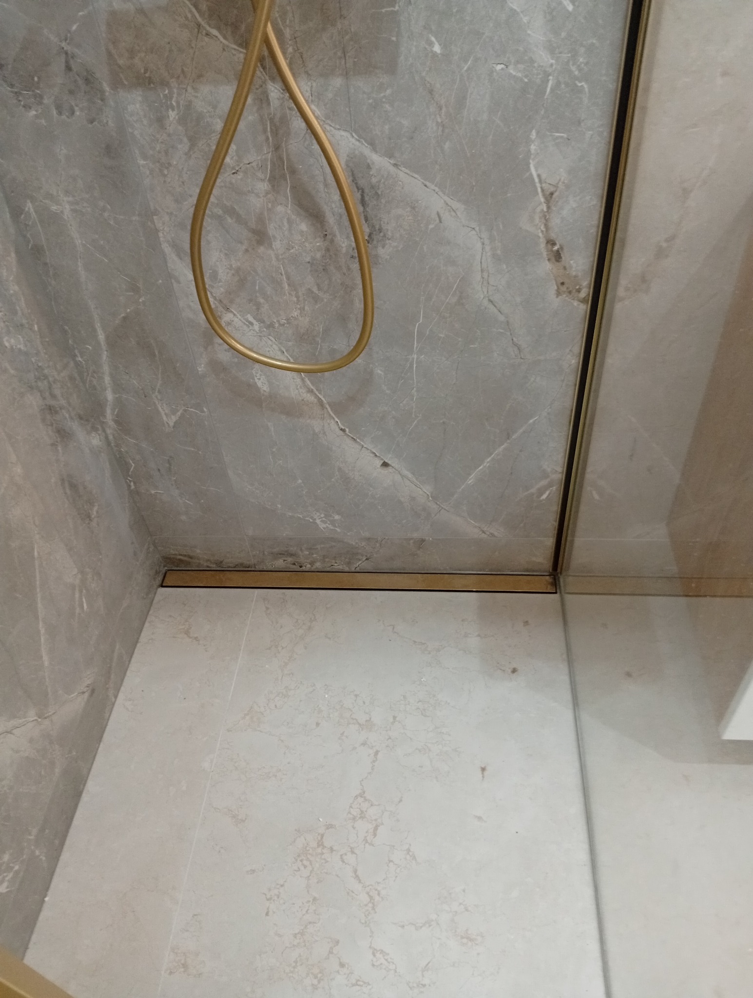 Wykończona łazienka: szare płytki imitujące marmur na ścianach i jasne na podłodze. Widoczny złoty odpływ liniowy i fragment kabiny prysznicowej z złotymi profilami.