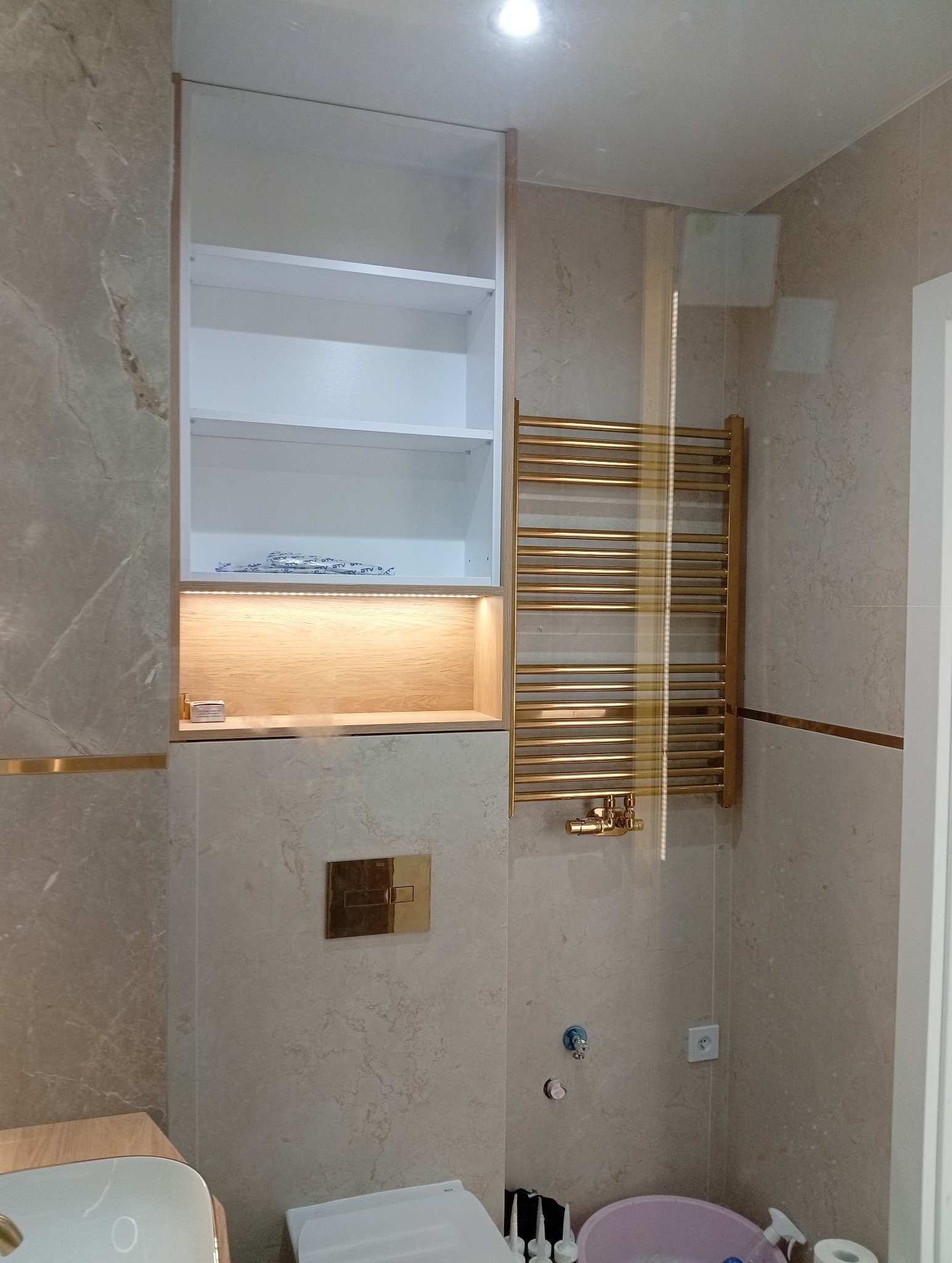Wykończona łazienka z marmurowymi płytkami, złotym grzejnikiem i zabudową szafki z podświetleniem LED. Widoczne elementy armatury i akcesoria łazienkowe.