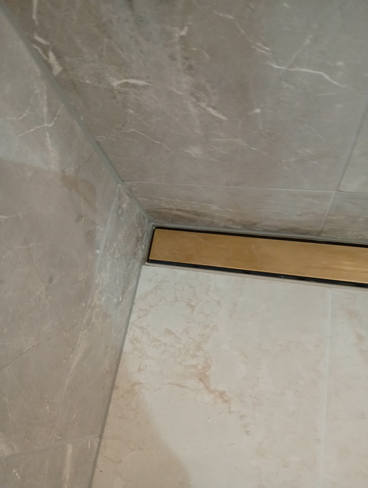 Wykończona łazienka z marmurowymi płytkami i złotym odpływem liniowym w rogu. Detal wykończenia pod kątem, ukazujący precyzję ułożenia płytek.