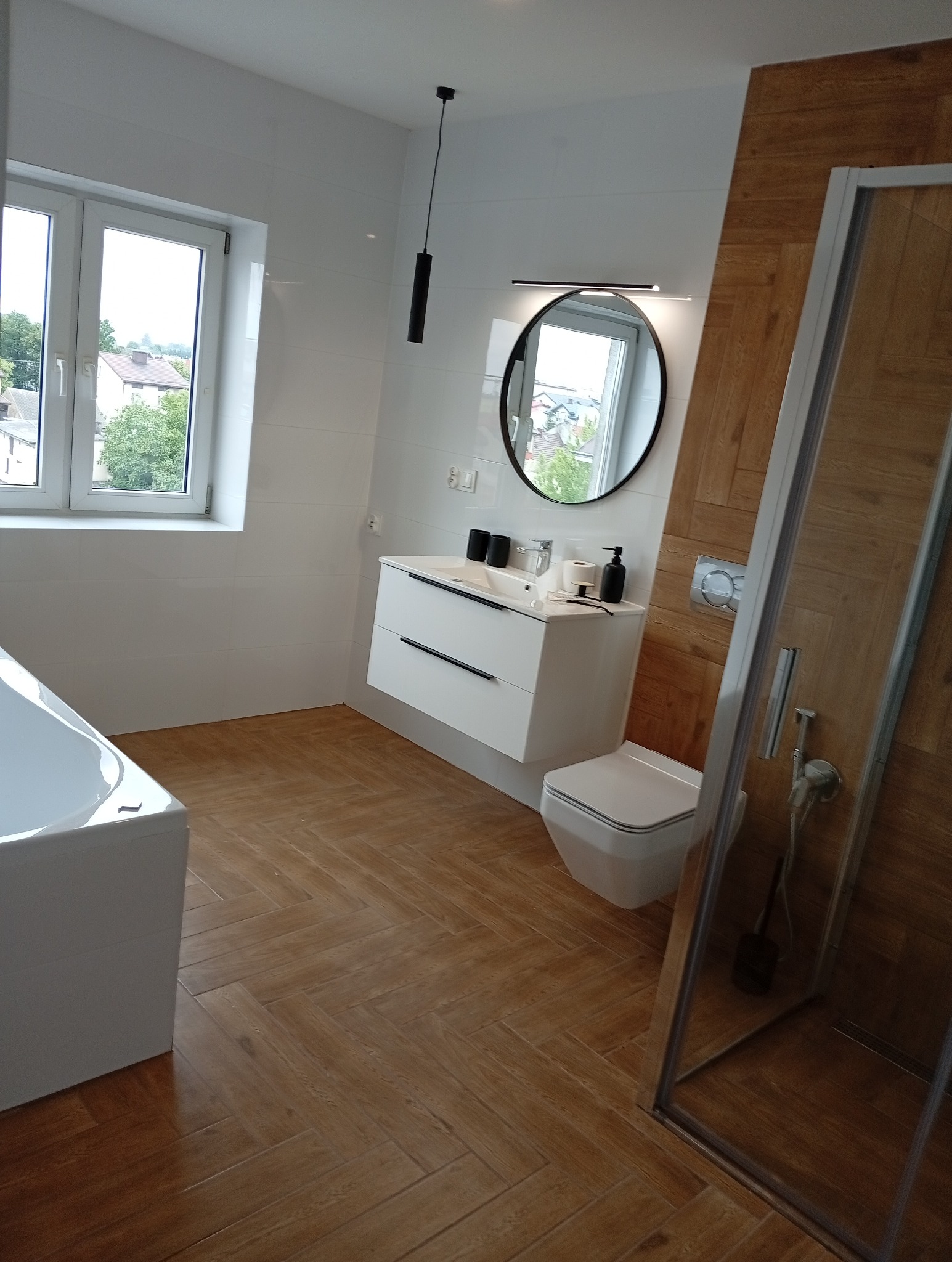 Nowoczesna łazienka z drewnopodobną podłogą ułożoną w jodełkę, białą armaturą i okrągłym lustrem. Widok na okno i zabudowę prysznicową.