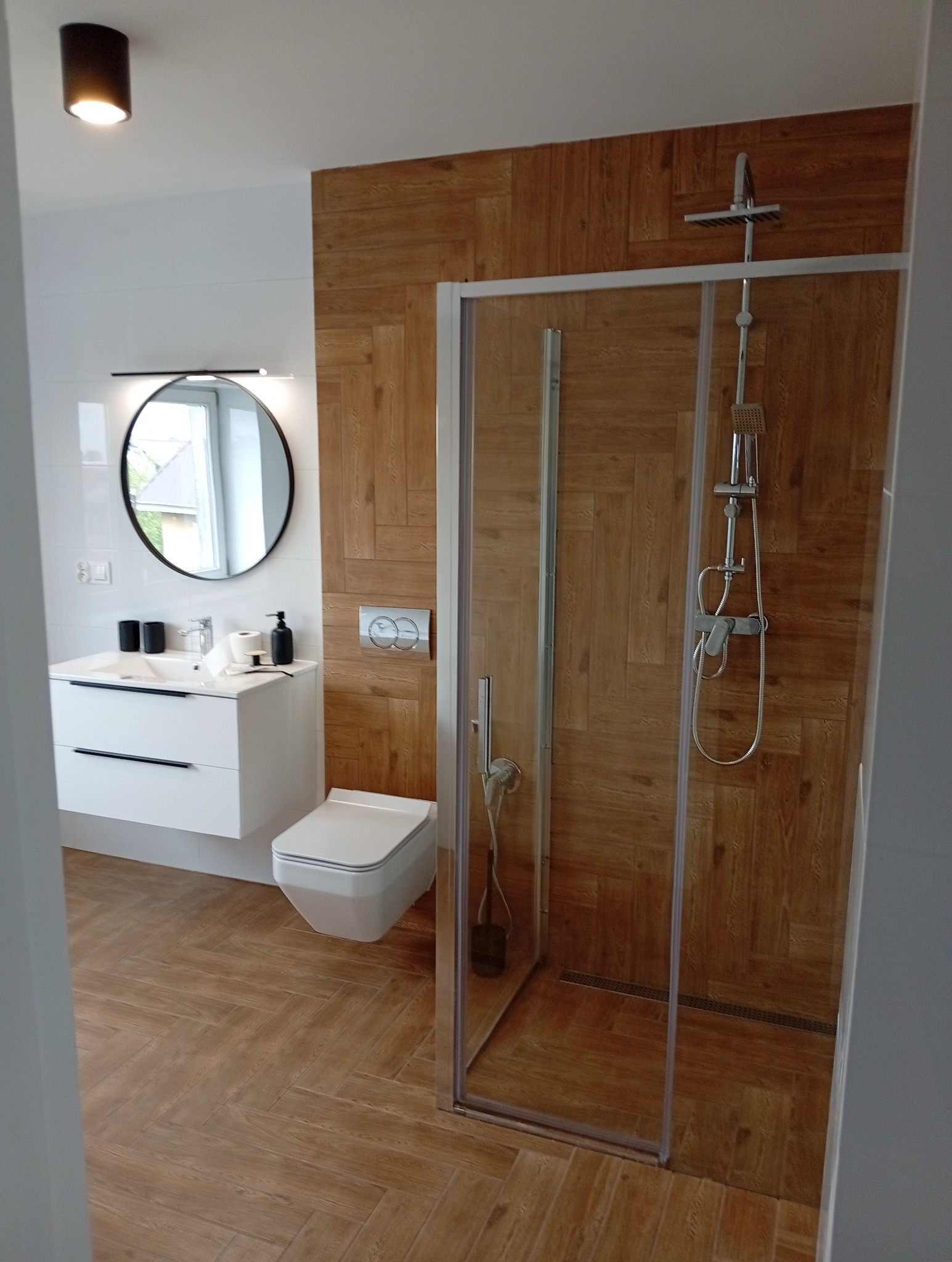 Nowoczesna łazienka z kabiną prysznicową obłożoną drewnopodobną płytką, białą umywalką z szafką i okrągłym lustrem. Uwagę przykuwają płytki ułożone w jodełkę.