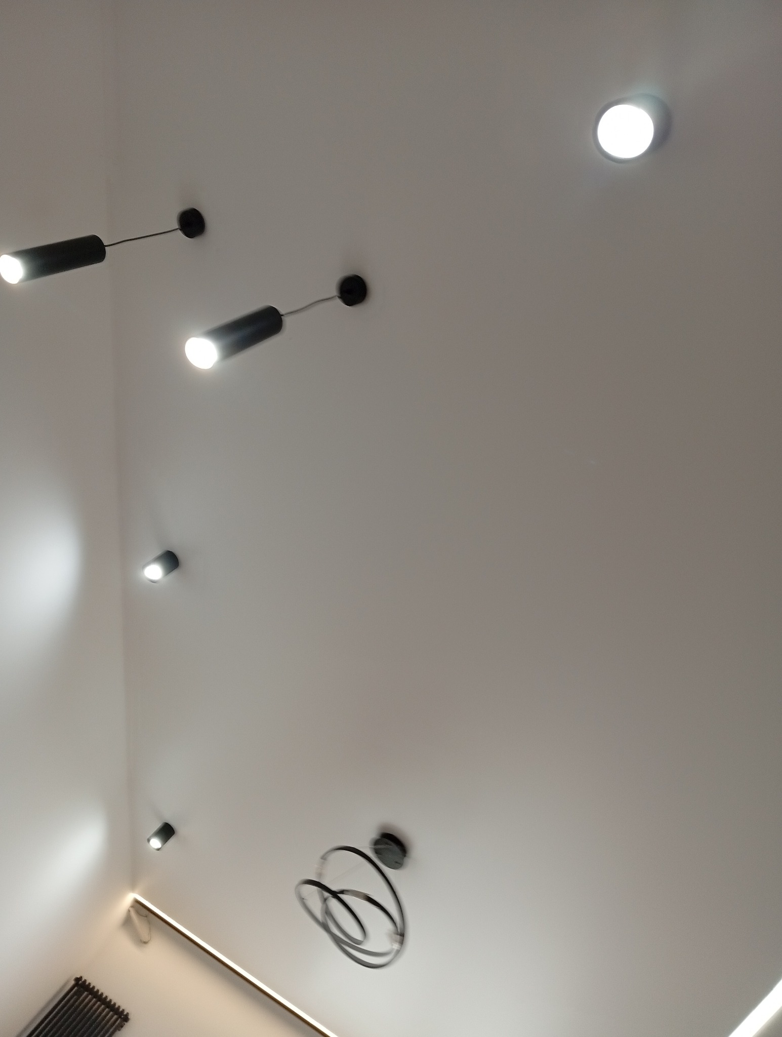 Biały sufit z zamontowanymi punktowymi czarnymi lampami oraz nowoczesnym wiszącym oświetleniem. Widoczny fragment listwy LED przy ścianie.
