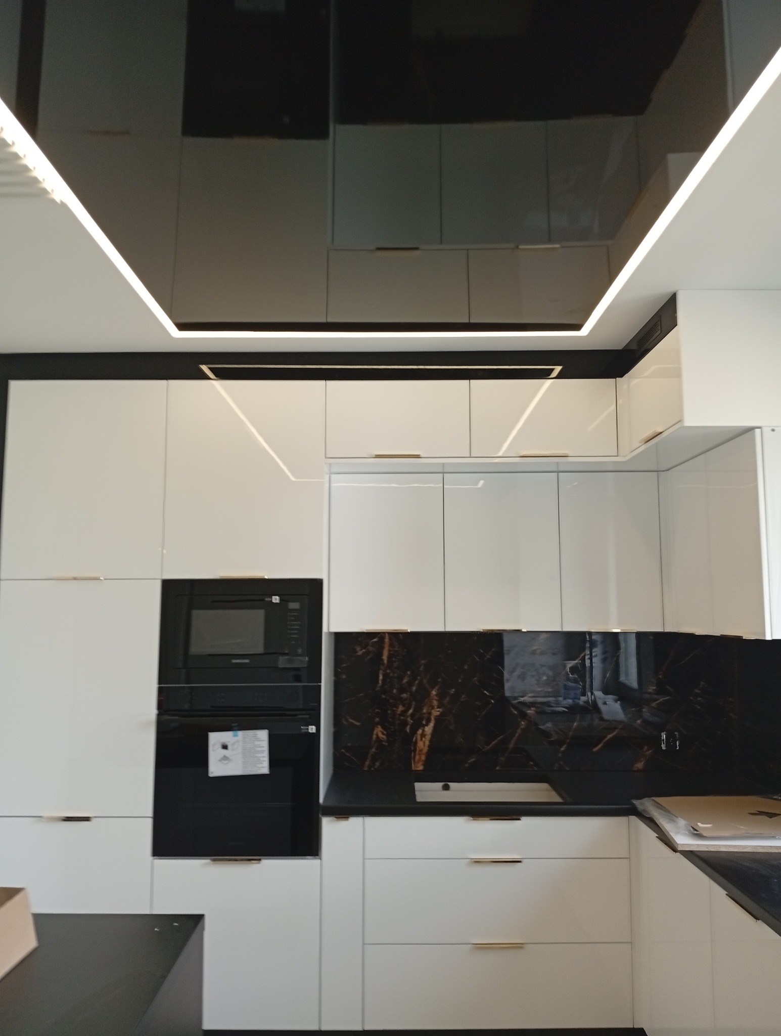 Nowoczesna kuchnia z białymi szafkami, czarnym blatem i marmurowym backsplashiem. Podwieszany sufit z oświetleniem LED podkreśla elegancki design.