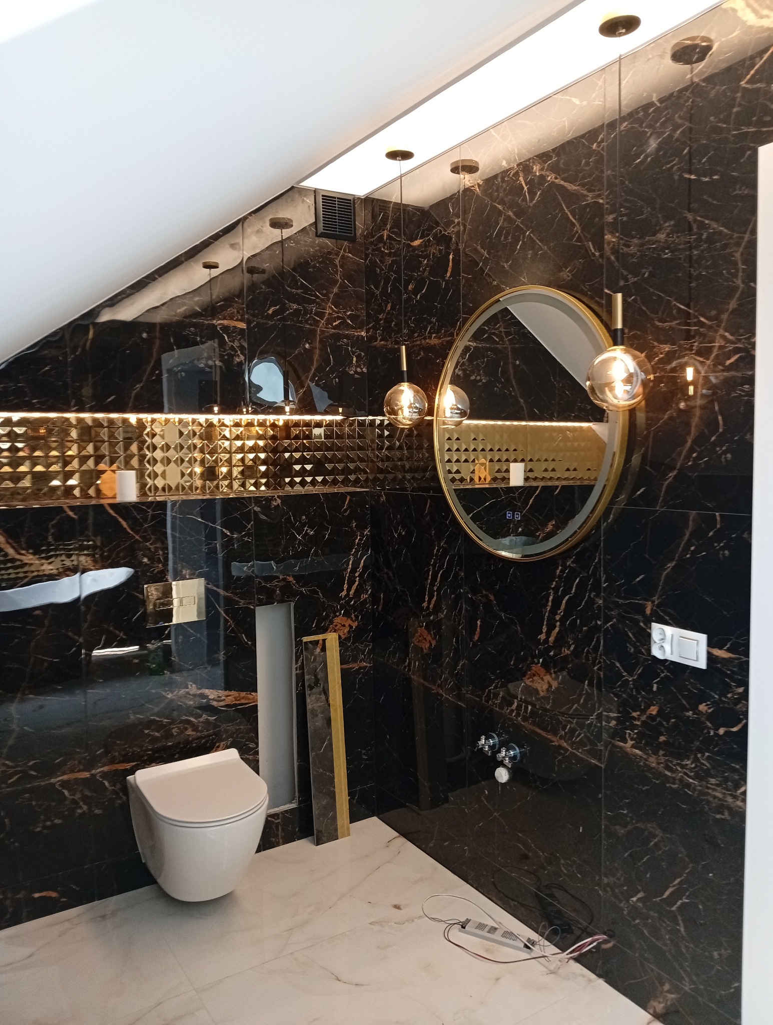 Luksusowa łazienka na poddaszu: czarne marmurowe płytki z złotymi żyłami, okrągłe lustro z podświetleniem i wisząca toaleta. Nowoczesny design z akcentem glamour.