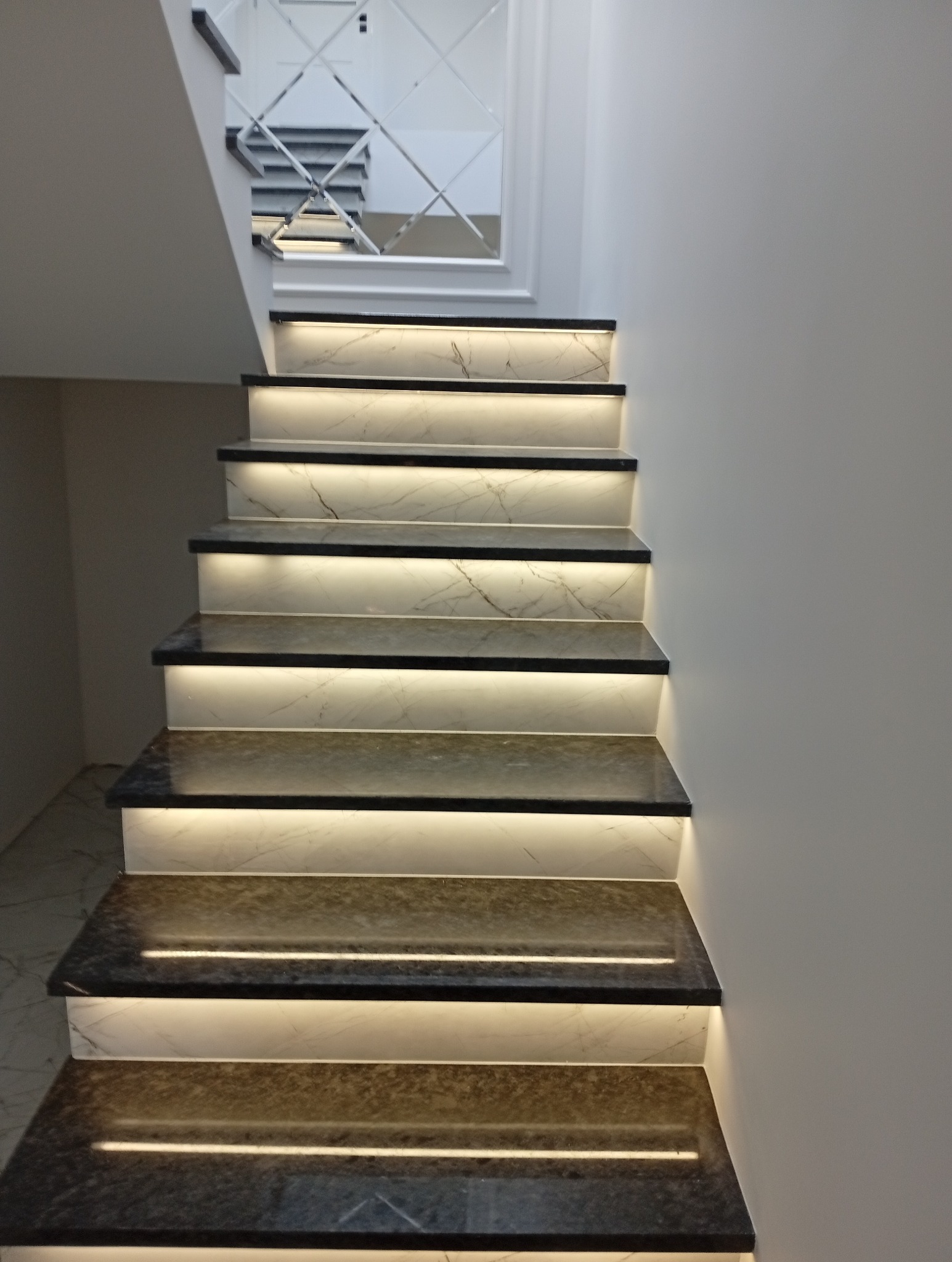 Schody z marmurowymi stopniami i podświetleniem LED, wykończone lustrem na ścianie. Nowoczesny design w jasnej tonacji. Efektowne oświetlenie podkreśla detale.