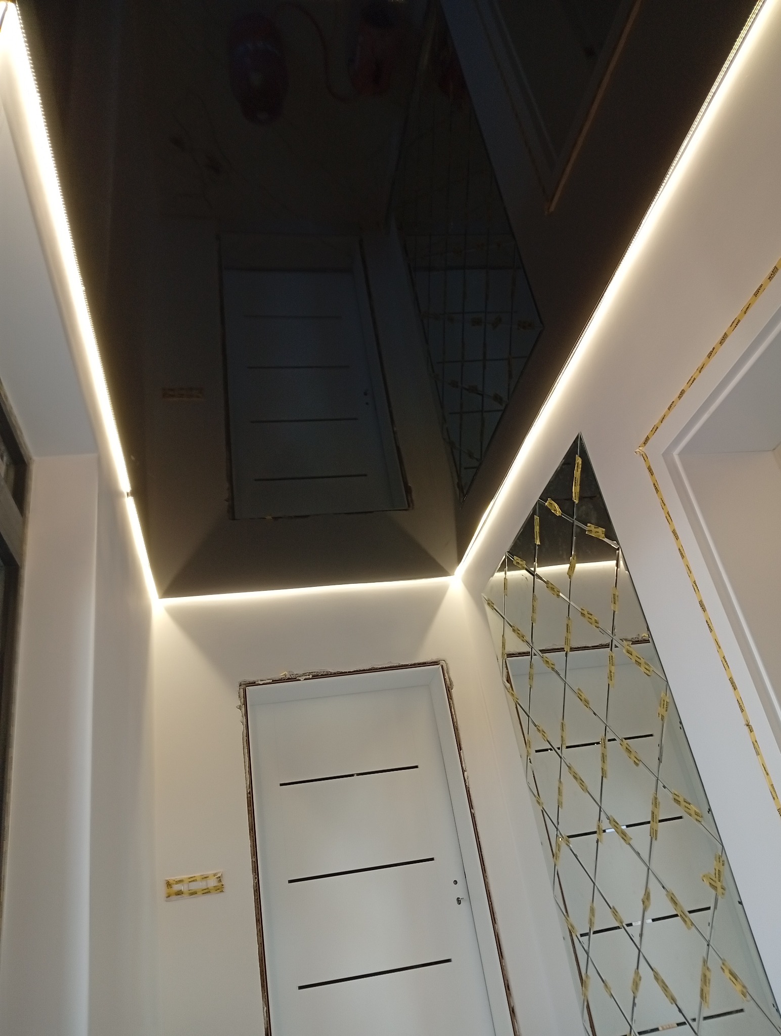 Błyszczący, czarny sufit napinany z oświetleniem LED wzdłuż krawędzi. Na ścianie dekoracyjne lustro w kształcie rombów. Białe drzwi z poziomymi wgłębieniami.