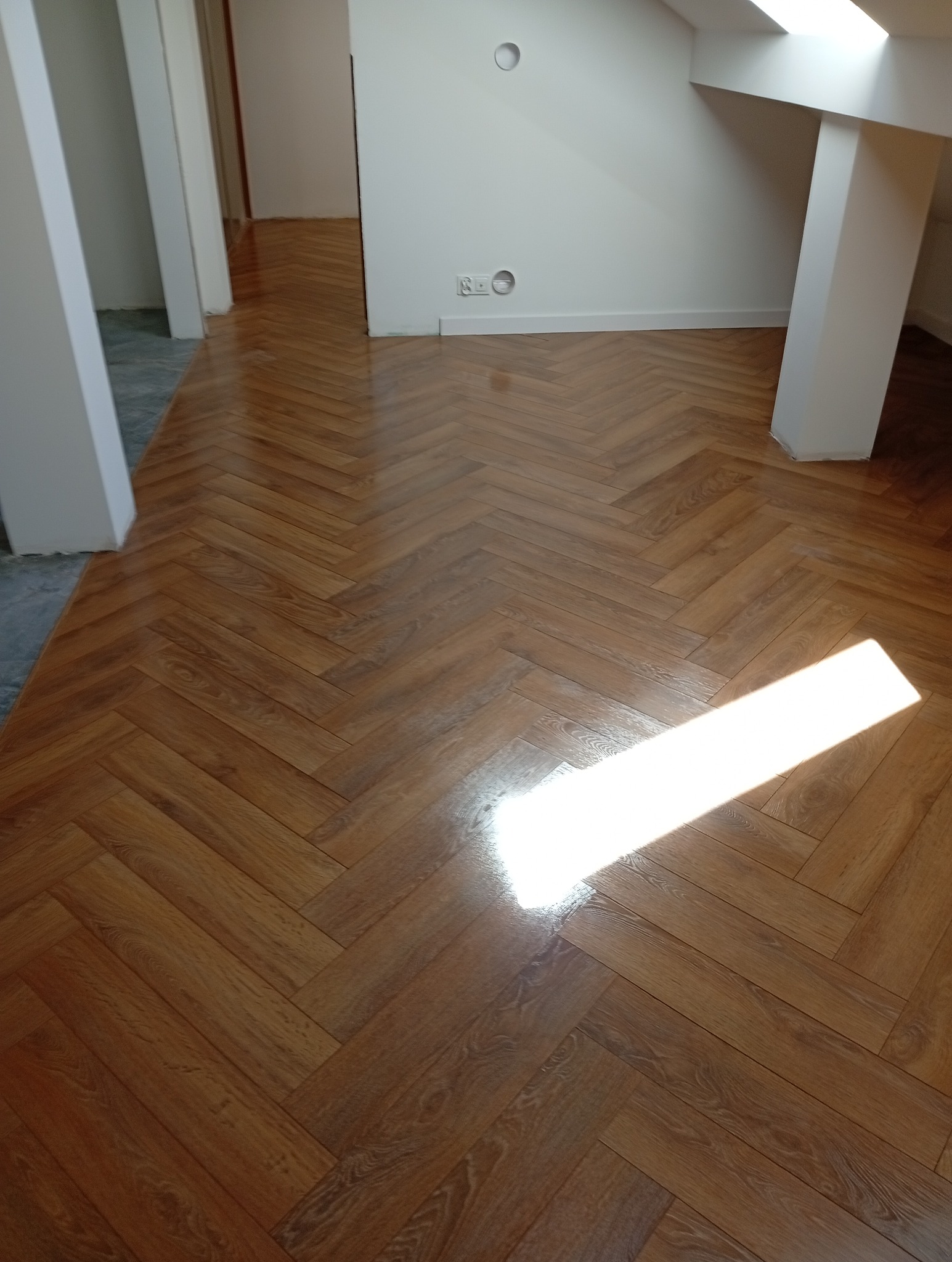 Parkiet ułożony w jodełkę w jasnym wnętrzu z białymi ścianami i filarami. Na podłodze widoczny blask światła wpadającego przez okno dachowe.