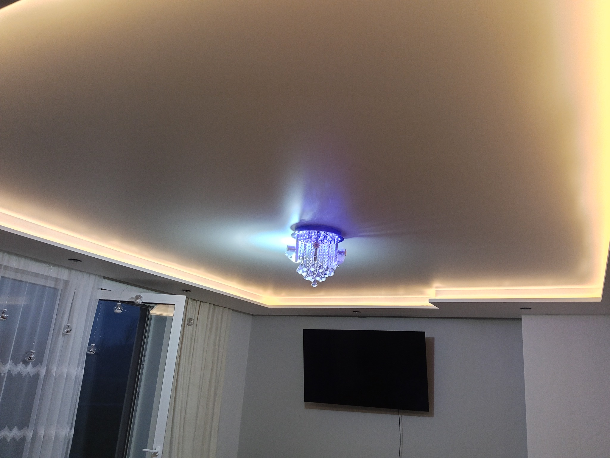 Biały sufit napinany z oświetleniem LED i kryształowym żyrandolem, widok z dołu. W tle okno z firankami i telewizor na ścianie.