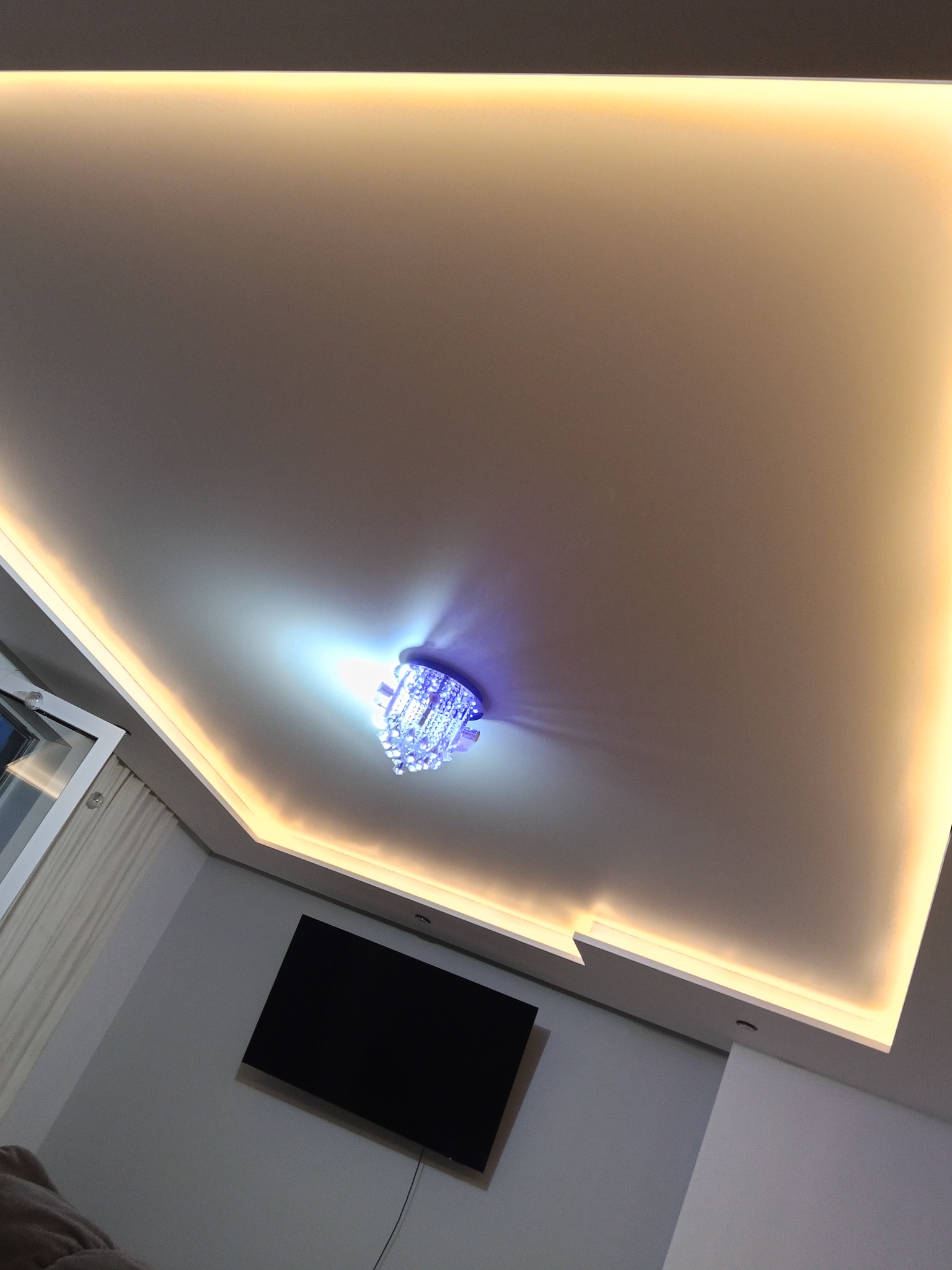 Biały sufit podwieszany z oświetleniem LED i kryształowym żyrandolem, widok z dołu. Na ścianie telewizor. Nowoczesne wykończenie wnętrza.