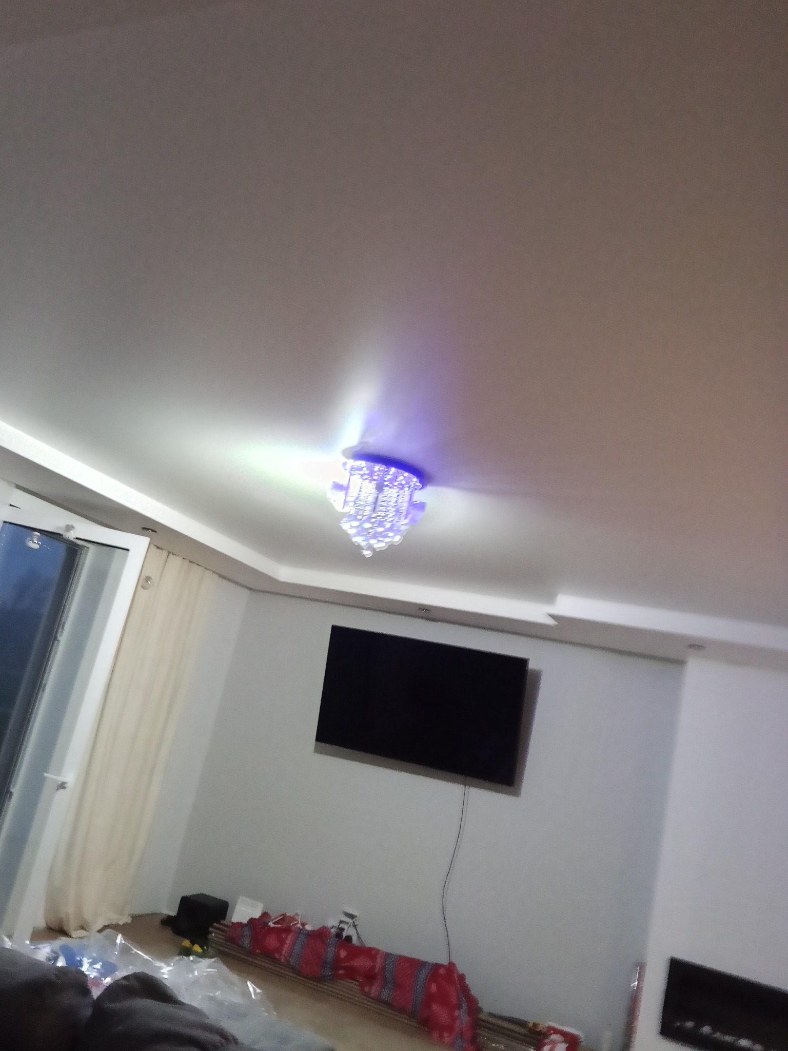 Biały sufit podwieszany z ozdobnym, kryształowym żyrandolem z niebieską poświatą, na tle ściany z telewizorem i oknem z zasłonami. Wykończenie wnętrz.