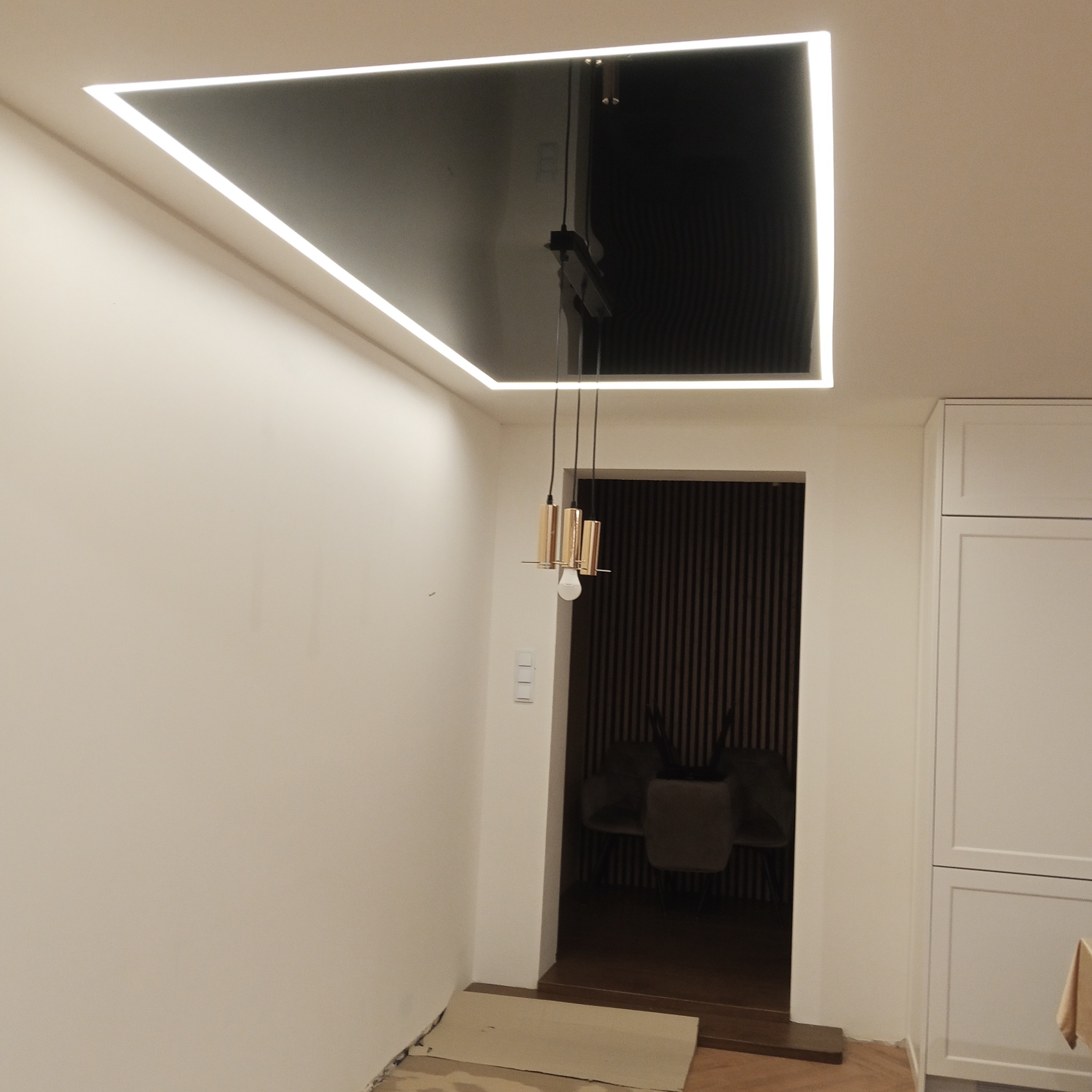 Czarny sufit napinany z oświetleniem LED w kształcie prostokąta, wisząca lampa z trzema złotymi cylindrami. Widok na jadalnię z krzesłami i ścianą w pionowe pasy.