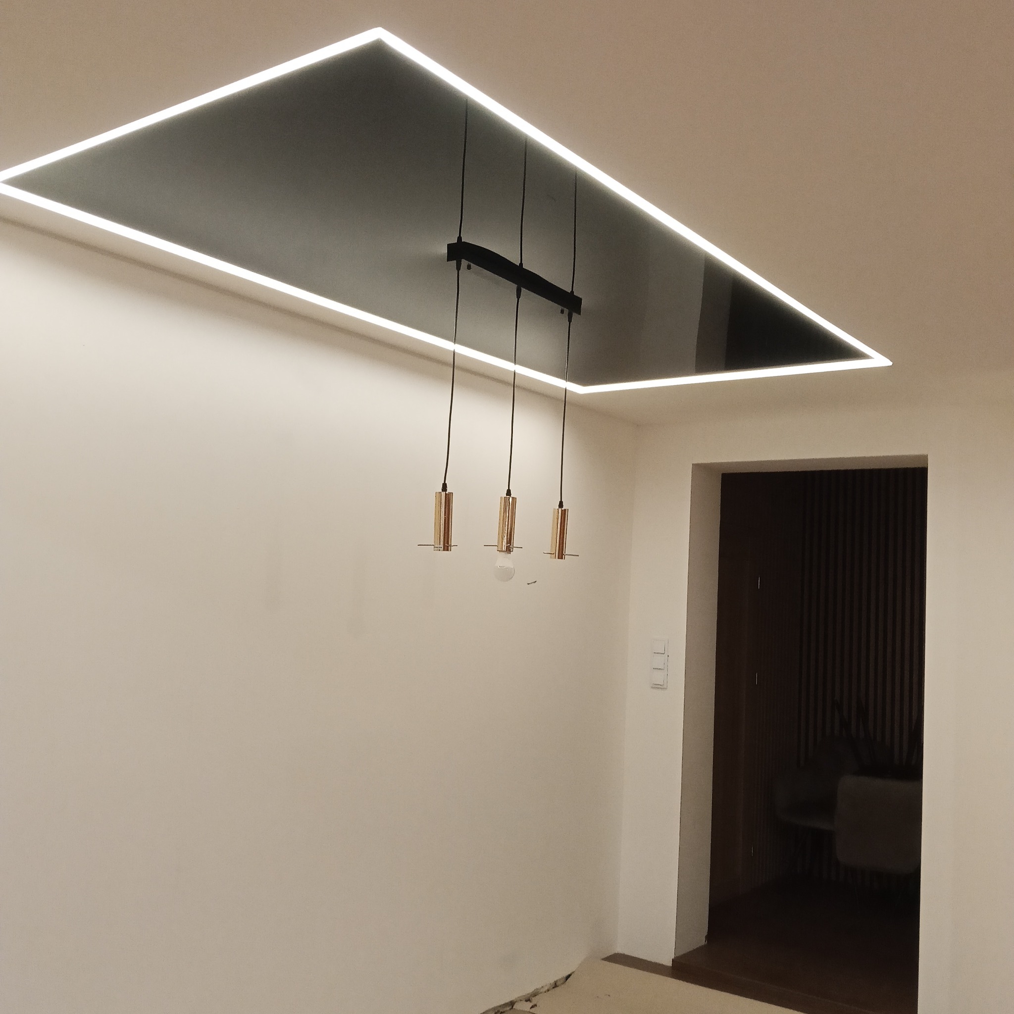 Nowoczesny sufit podwieszany z czarną, błyszczącą powierzchnią i obwodowym oświetleniem LED. Wiszące lampy z elementami w kolorze miedzi. Minimalistyczne wnętrze.