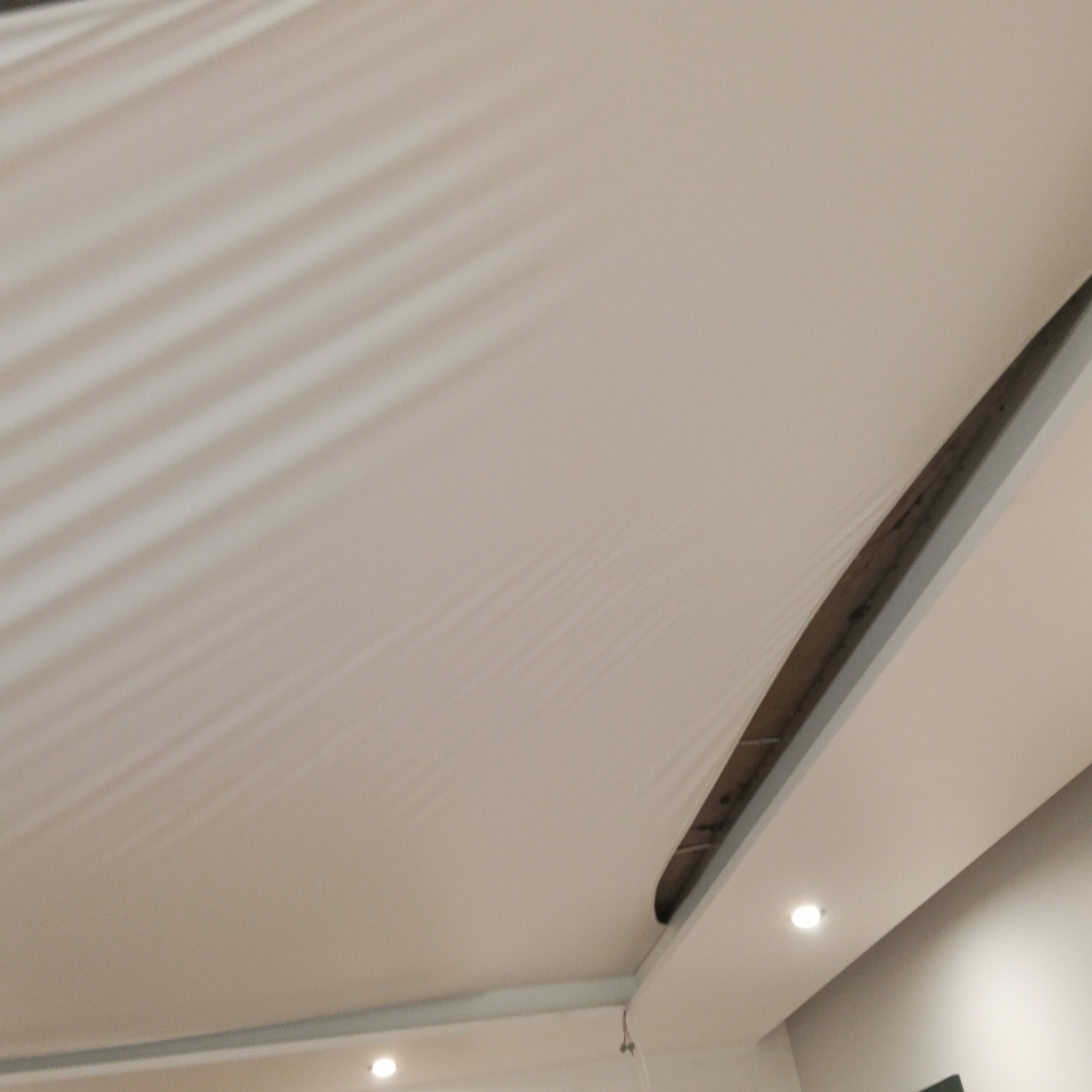 Biały sufit napinany z delikatnym wzorem cieni, widoczny fragment konstrukcji montażowej i oświetlenie punktowe. Ujęcie z dołu, podkreślające gładką powierzchnię.