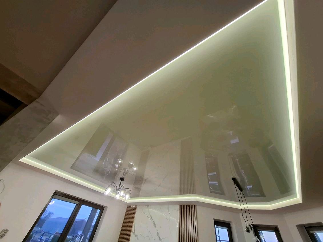 Błyszczący, beżowy sufit napinany z oświetleniem LED wzdłuż krawędzi, odbijający światło z okien i żyrandola. Widoczne elementy dekoracyjne na ścianie.