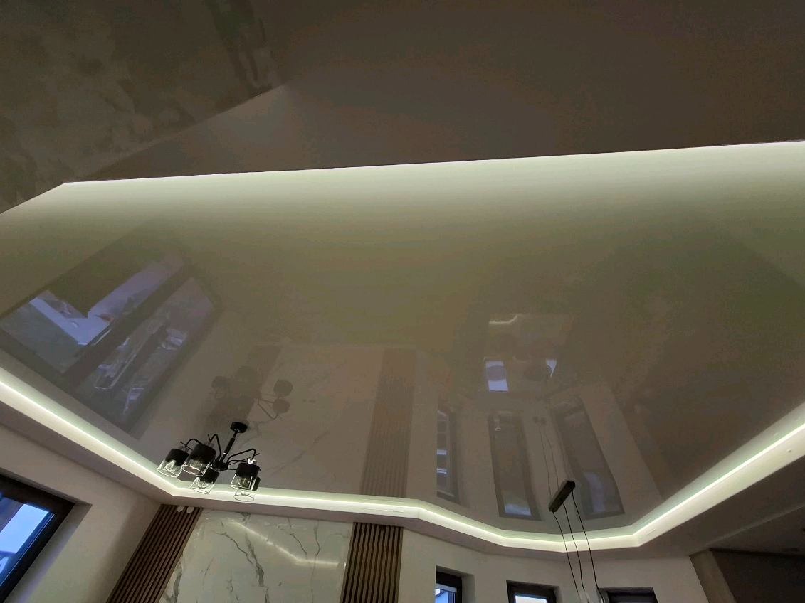 Błyszczący, beżowy sufit napinany z oświetleniem LED, odbijający elementy wystroju wnętrza, w tym żyrandol i okna. Widok z dołu.