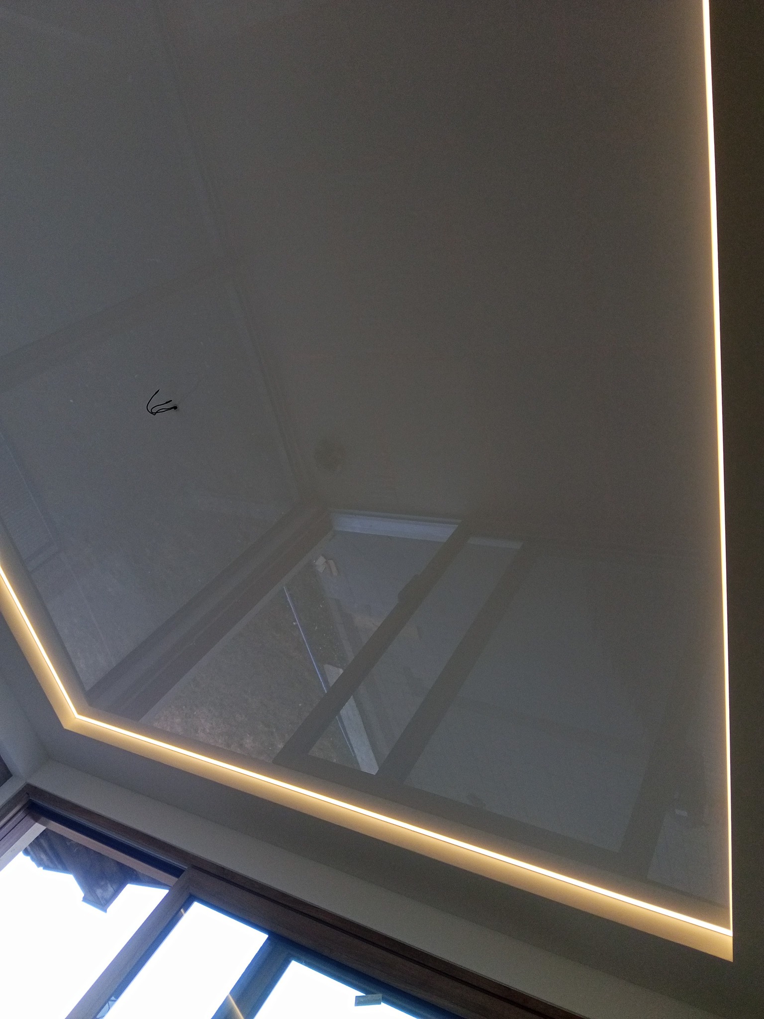 Błyszczący, szary sufit napinany z oświetleniem LED wzdłuż krawędzi, widok z dołu, okno w tle. Widoczne kable wystające z sufitu.