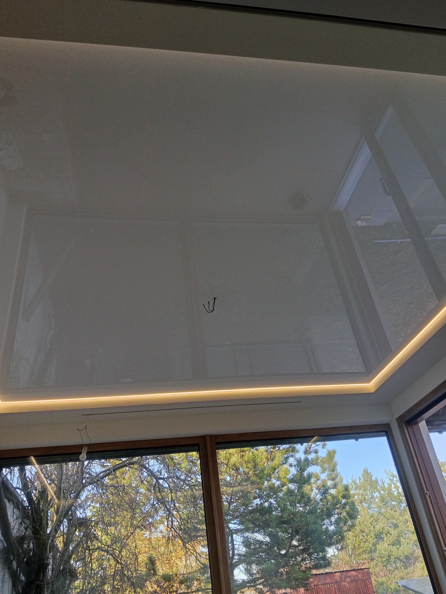 Biały sufit napinany z oświetleniem LED wzdłuż krawędzi, widok z dołu na okno z widokiem na drzewa i niebo. Widoczne elementy montażowe.