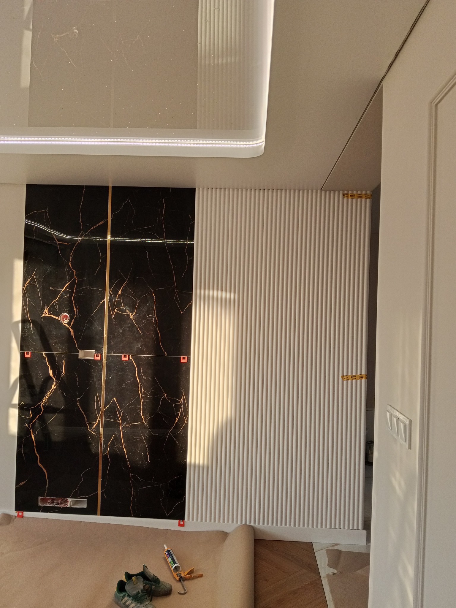 Ściana z czarnych płyt imitujących marmur z złotymi żyłami i białe panele ścienne z pionowymi żłobieniami. Widoczne narzędzia i buty na podłodze.