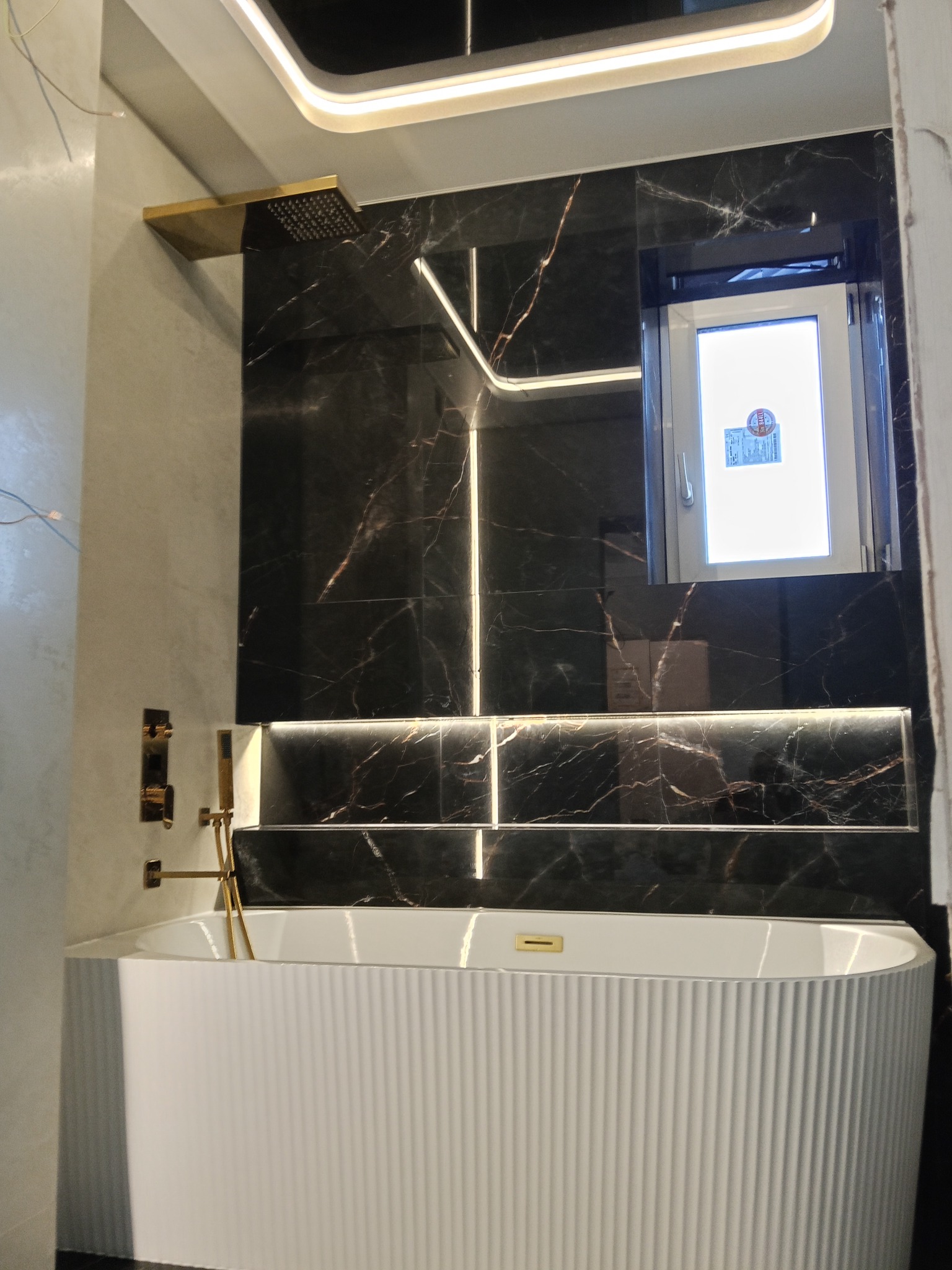 Nowoczesna łazienka z czarną marmurową ścianą, podświetleniem LED i białą wanną z żłobieniami. Złota armatura i deszczownica dodają elegancji.