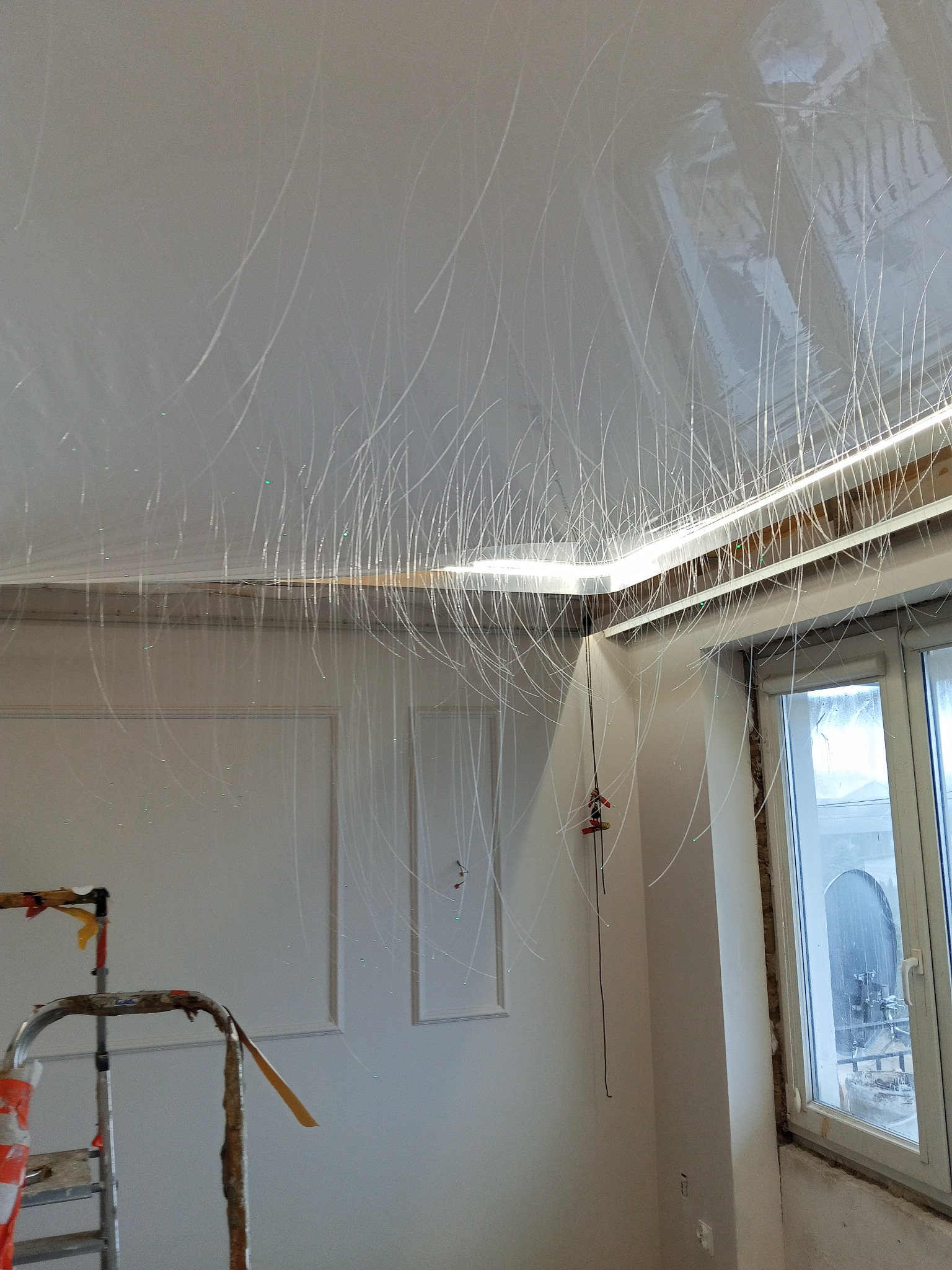Błyszczący, biały sufit napinany z delikatnymi, srebrzystymi wzorami, częściowo widoczna drabina i okno, wnętrze w trakcie wykończenia.