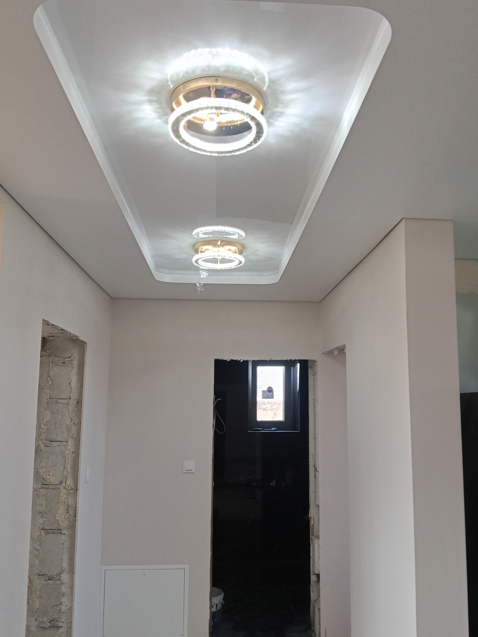 Biały sufit napinany z dwoma okrągłymi lampami LED w kształcie pierścieni, widok z dołu w trakcie wykańczania wnętrza, z widocznymi otworami drzwiowymi.