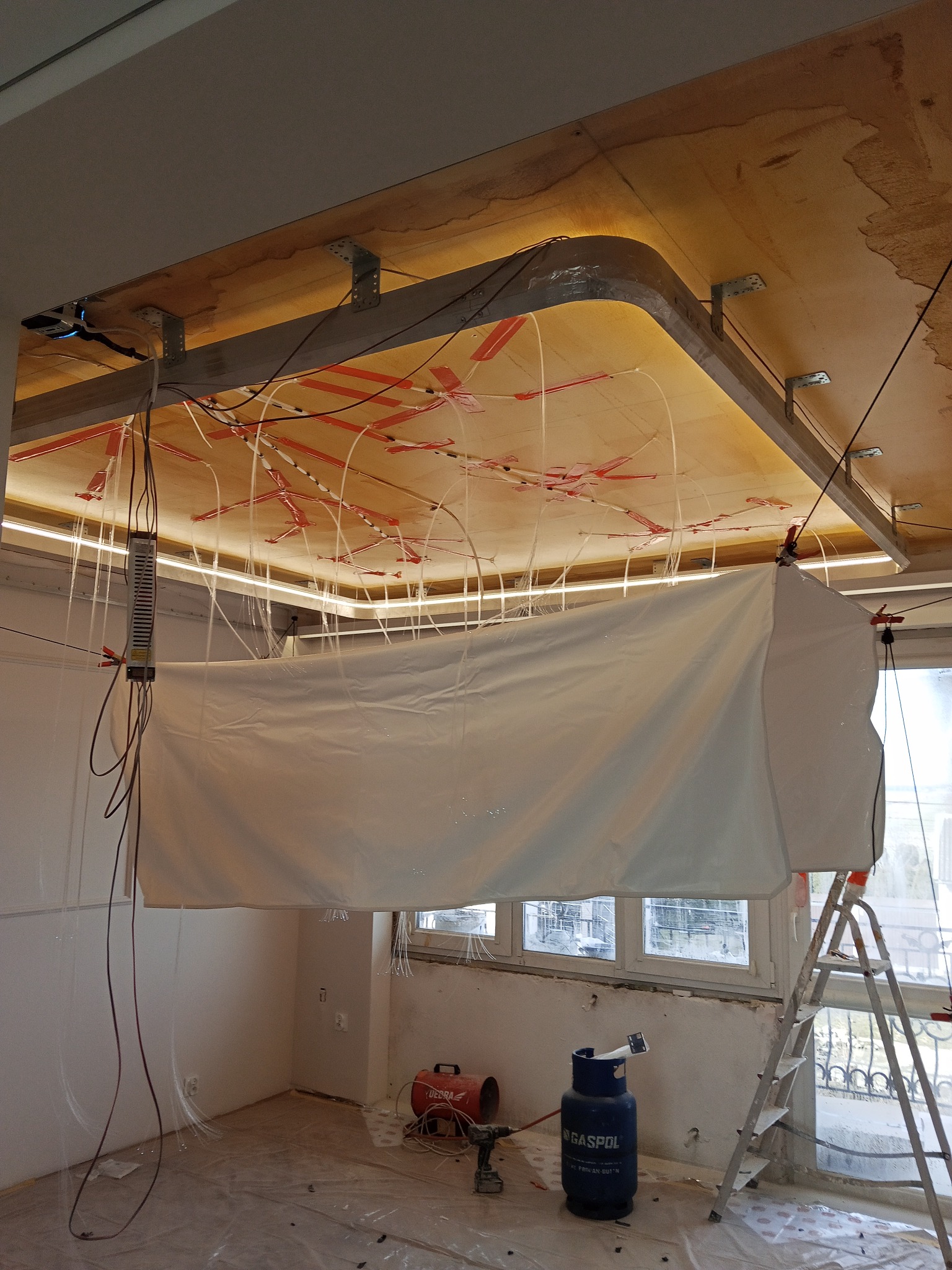 Montaż sufitu napinanego: widoczne ramy, instalacja oświetleniowa i folia ochronna. Przygotowania do montażu, butla gazowa i drabina w pomieszczeniu.