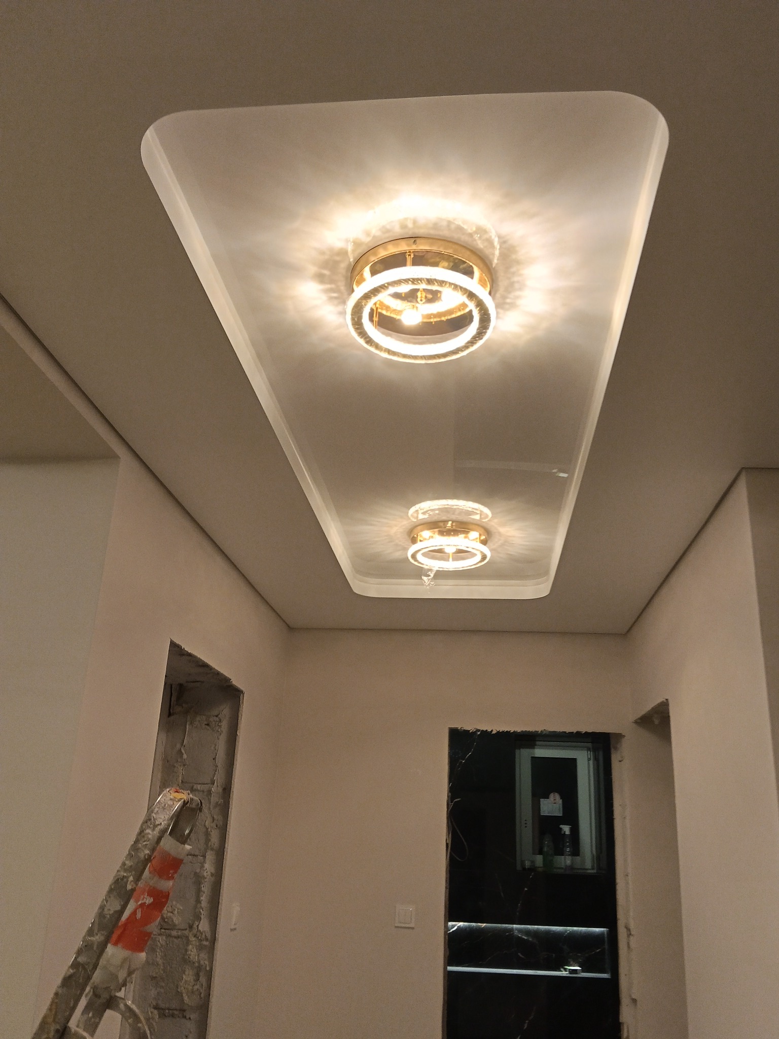 Biały sufit podwieszany z dwoma okrągłymi lampami LED w kształcie pierścieni, widok z dołu, fragment drabiny w rogu, drzwi w tle.