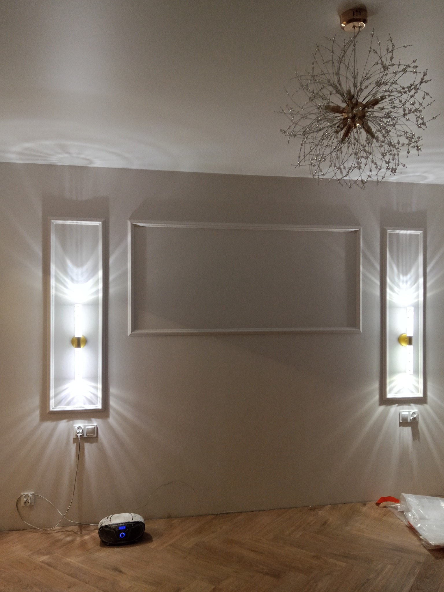 Eleganckie wykończenie wnętrza: dekoracyjne listwy ścienne, symetryczne kinkiety z ciepłym światłem i designerski żyrandol w kształcie gwiazdy nad parkietem.