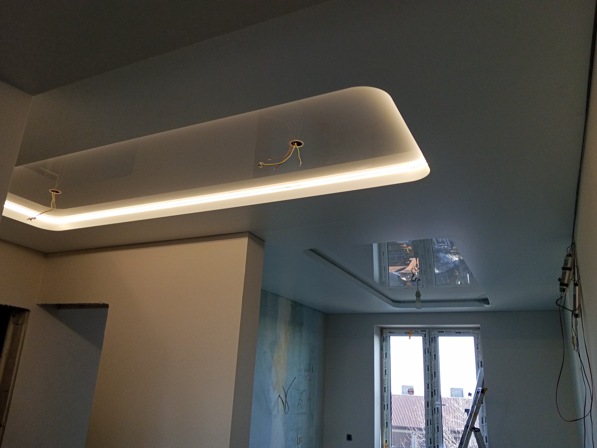 Nowoczesny sufit napinany z podświetleniem LED w kształcie prostokąta, widok z dołu. W tle okno i drabina. Częściowo wykończone wnętrze.