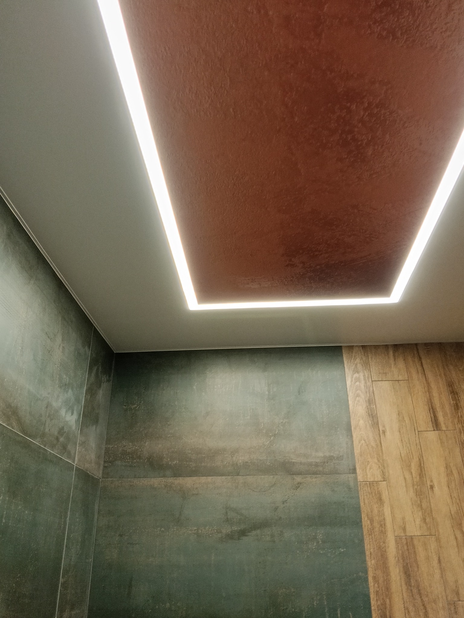 Nowoczesny sufit z podświetleniem LED w kształcie prostokąta, brązowa faktura sufitu, ściany wykończone płytkami w odcieniach zieleni i drewnopodobnymi panelami.