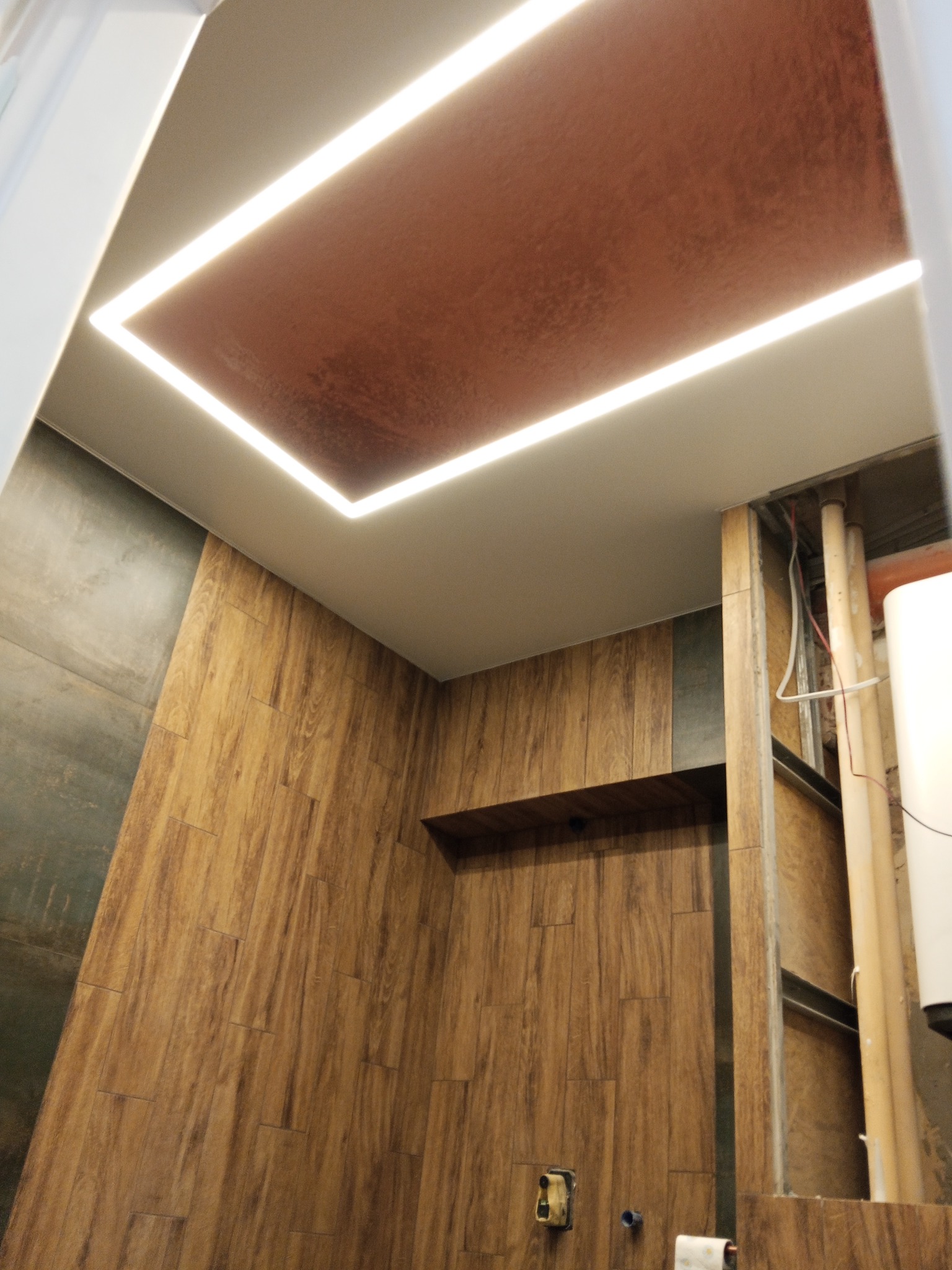 Nowoczesny sufit napinany z oświetleniem LED w łazience w Chamsku. Brązowa powierzchnia sufitu i drewnopodobne płytki na ścianach tworzą ciepły klimat.